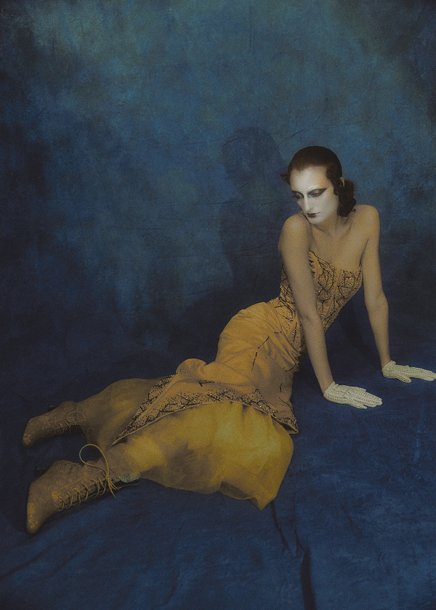 Seated figure in a gold dress against a painted blue backdrop